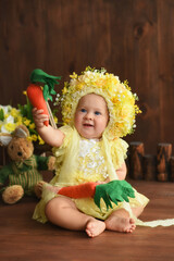 Portrait of a little girl in a yellow suit. Girl 7 months old sitting. Little child in a hat made of flowers. Little child holding a carrot in his hands