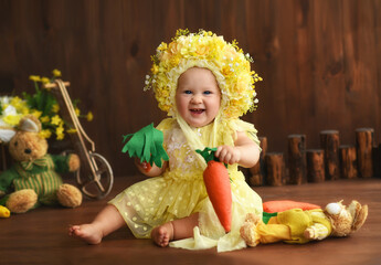 Portrait of a little girl in a yellow suit. Girl 7 months old sitting. Little child in a hat made of flowers. Little child holding a carrot in his hands