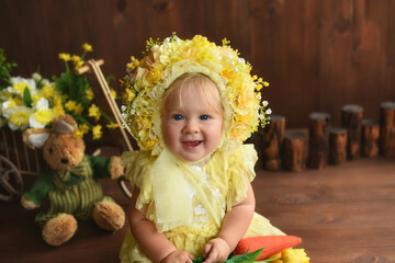 Portrait of a little girl in a yellow suit. Girl 7 months old sitting. Little child in a hat made of flowers. Little child holding a carrot in his hands