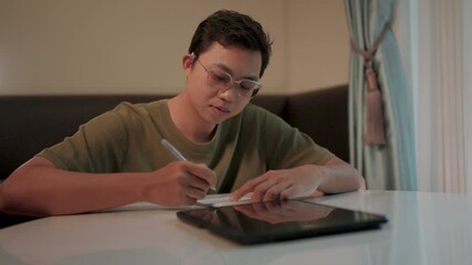 Focused on the task a young man with glasses concentrates intently on writing notes, his stylus poised over a tablet, showcasing a dedicated and determined approach to work or study. 
