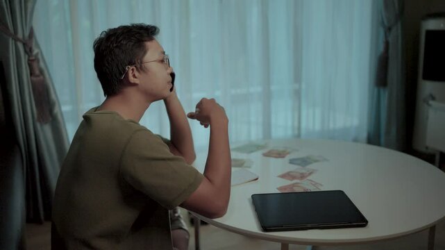 Late Night Decision: A pensive individual sits at a table, illuminated by the soft glow of a window, engaged in a phone call while surrounded by scattered papers and a tablet.