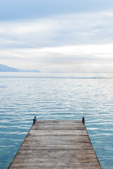 Naklejka premium Tranquil scene featuring a wooden pier extending into a calm blue lake, with distant mountains and a cloudy sky creating a peaceful atmosphere.
