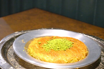 Close-up of traditional Turkish syrupy dessert kunefe. Kunefe with pistachios. Kadayif desserts. Lemon and brewed tea in a glass cup. Presentation on a copper plate. Turkish desserts. Food industry.