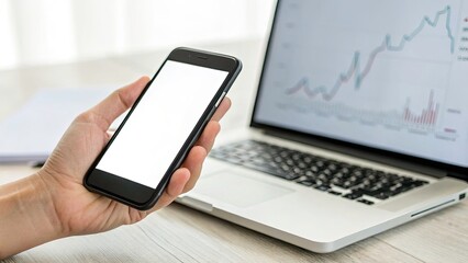 Minimalist Close-up of Hand Holding Mobile Phone with Blank Display, Laptop and Blurred Graph Background, Showcasing Technology in Business and Finance Concepts