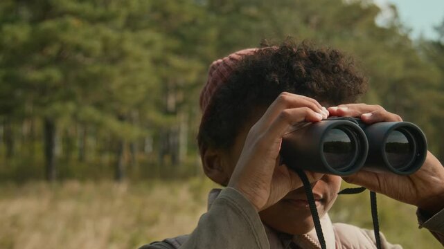 Close-up shot of African American teenage kid looking through binoculars at river landscape during autumn hike with family