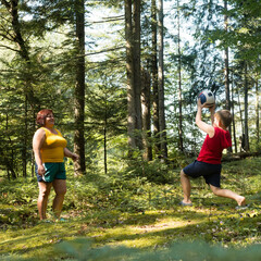 An adult woman and a young boy enjoy a game of catch in a lush forest setting. Sunlight filters through the trees, illuminating the scene.