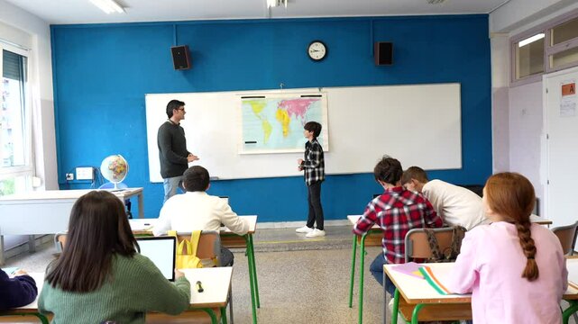 Student presenting geography lesson, explaining world map details to attentive classmates while standing near whiteboard in classroom setting