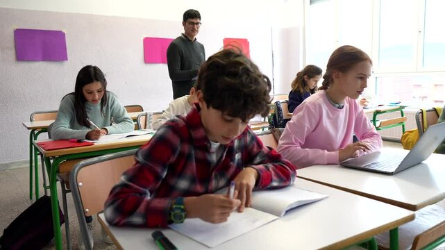 Young students are engaged in their studies, writing and using laptops in a brightly lit classroom, with a teacher overseeing the activities