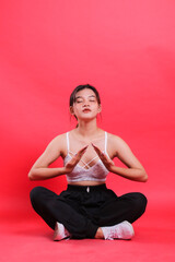 Fototapeta premium A young woman is sitting in a meditating pose with her eyes closed, wearing a white top, black pants, and white shoes, isolated on a red background