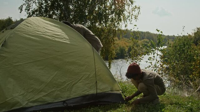 Tilt down view of gen alpha kid helping father to set up green rainfly when pitching tent on hill above river in early fall during family hike