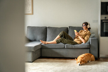 Happy smiling young student woman lying on couch, listening music in headphones on mobile phone...