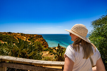Fototapeta premium Summer vacation in Lagos Portugal. Mature woman tourist in sunglasses and sun hat standing on viewpoint on cliffs during sightseeing Ponta da piedade in Algarve on beautiful sunny day. Back view
