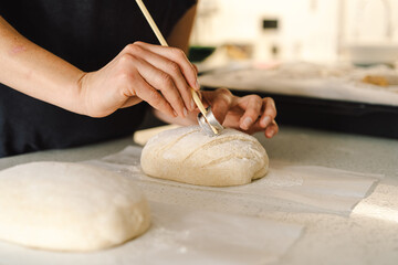 Woman hands skillfully score the surface of a round dough loaf, preparing sourdough bread for...