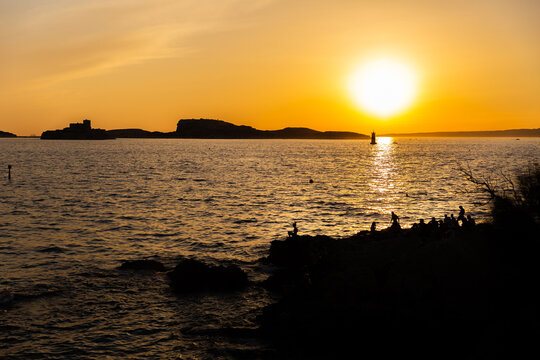 Coucher de soleil sur un paysage marin &agrave; Marseille, une &icirc;le en arri&egrave;re plan et ligne d'horizon avec dix personnes assises sur des rochers en premier plan