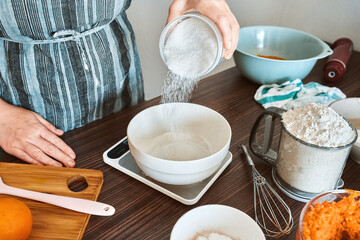 Woman cook in apron pouring sugar into bowl on kitchen scales to prepare carrot cake on kitchen table surrounded by other ingredients.