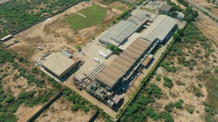 Aerial view capturing a large industrial factory in Karachi, Pakistan, featuring multiple buildings and a cricket field amidst arid land and sparse vegetation