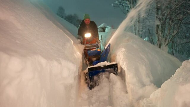 An adult man using snowblower machine to clear deep snow in heavy snowfall at night