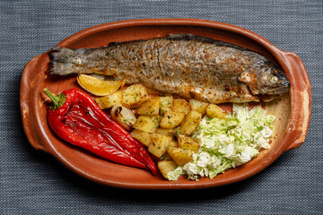 Tasty baked trout fish with red pepper, potatoes and Chinese cabbage salad in a clay dish on the table, closeup, top view
