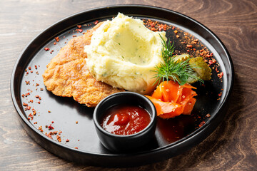 A delicious plate featuring a crispy fried cutlet, creamy mashed potatoes, fresh vegetables, and a small bowl of ketchup, served on a black plate with a wooden background