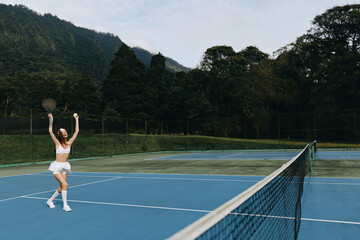 Joyful female tennis player celebrating on a blue court, wearing a stylish white outfit, with a scenic mountain backdrop showcasing vibrant green trees