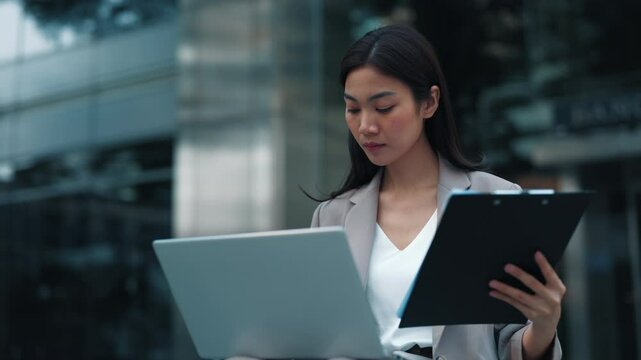 Portrait of Thai woman entering data from work papers into laptop sitting on city street.