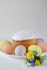 An Easter cake with white icing and pastry beads stands on a white plate. Around it are painted eggs in pastel shades and a small bouquet of spring flowers.
