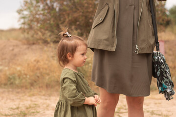 Little cute girl in green dress standing next to mother's skirt in summer outdoors. Concept of...