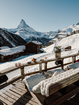 Cozy alpine terrace with a breathtaking view of the Matterhorn. A rustic wooden bench covered in soft fur, warm drinks, and mountain chalets create the perfect winter retreat in Zermatt, Switzerland