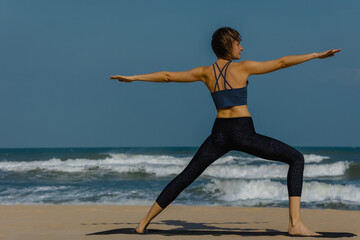 Fototapeta premium Female adult practicing yoga on beach with ocean waves under blue sky
