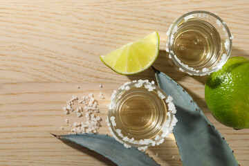 Tequila shots with salt, limes and agave leaves on wooden table, flat lay. Space for text