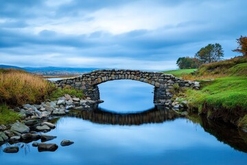 Fototapeta premium Ancient Stone Bridge Arching Over Slow Moving River Landscape