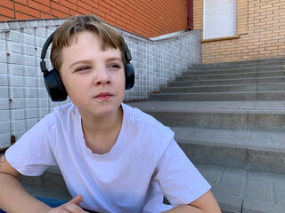 A boy in a white shirt sits on concrete steps, wearing headphones. He gazes into the distance,...