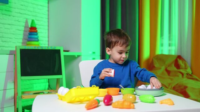 Active toddler wearing blue sweater sits at desk playing with toys. Happy kid having a game with plastic eggs, crockery and vegetables.