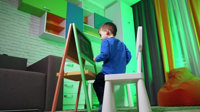 Small kid sits down on the chair in front of the blackboard. Low angle view at the toddler drawing with chalk and cleaning the picture.