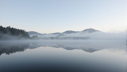 Fototapeta premium Mysterious mist shrouds a tranquil lake at dawn with mountains fading in the ethereal fog