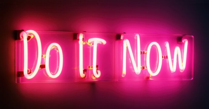 Glowing neon sign displaying Do It Now in bold pink letters, mounted on a dark background, radiating urgency, motivation, and determination to take immediate action