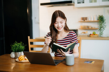 Asian woman working remotely on a laptop and tablet in a cozy kitchen workspace. Perfect for education
