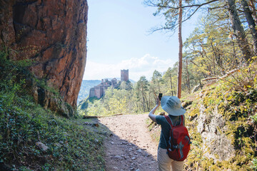 A woman wearing a sun hat and a red backpack takes a photo of The Three Castles of Eguisheim while hiking through a scenic forest trail with rugged cliffs and lush greenery.