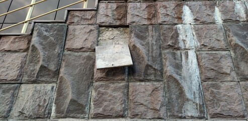 Wall of the building. Textured wall covered with stone tiles. The tiles are uneven and imitate real stone. One tile has fallen off and is hanging on a metal fastener cement wall is visible behind it.