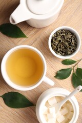 Refreshing green tea in cup, teapot, sugar bowl and leaves on wooden table, flat lay