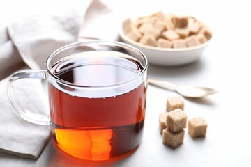 Aromatic black tea in cup and brown sugar cubes on white table, closeup