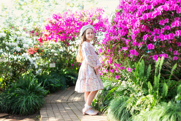 Naklejka premium Beautiful caucasian little girl in greenhouse among bush colorful azalea flowers. Portrait of a child girl wearing pink dress and wear beret in the botanical garden arbor among the blossom flowers. 