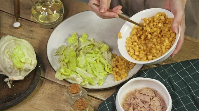 Iceberg lettuce or cabbage leaves are neatly arranged on plate, and chef adds canned corn kernels. Housewife prepares nutritious vegetable salad with seafood