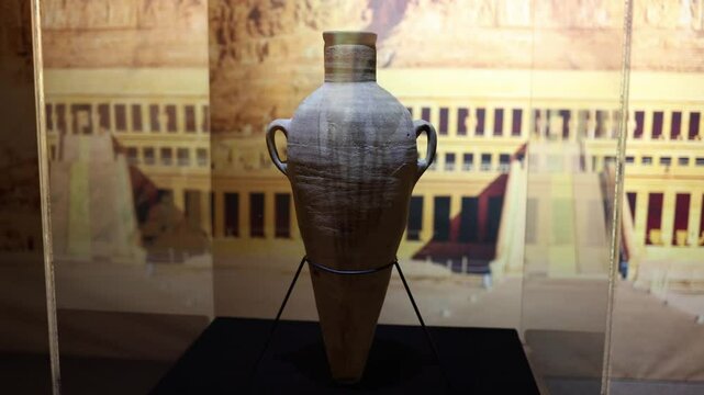 An ancient Egyptian amphora displayed in a museum, set against a backdrop of historical architecture. The well-preserved clay vessel reflects the craftsmanship of a lost civilization.