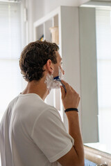 A vertical image of a young man carefully shaving his beard while standing in front of a bathroom mirror, focusing on personal grooming and daily routines.