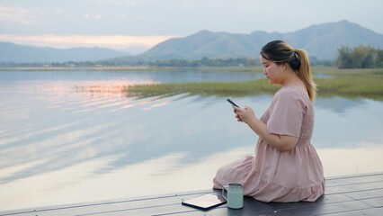 Woman Using Tablet by Lake at Sunset, Surrounded by Majestic Mountains. Digital nomad freelancer,...