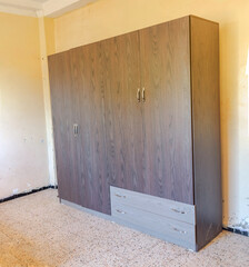 A new, modern dark brown wooden wardrobe with two gray drawers, four doors and silver handles, in a room with granite tiled flooring and a deteriorated wall with black baseboard stained with paint. 