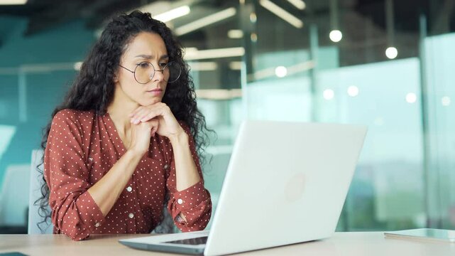Thoughtful serious young businesswoman working on laptop while sitting at a desk at a workplace in business office. Focused female manager works on a complex project, thinks about solving a problem