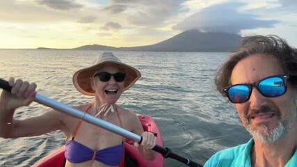 A mature man and woman share a joyful moment taking a selfie while kayaking in Lake Nicaragua during sunset. The backdrop features the stunning Conception volcano in Central America.