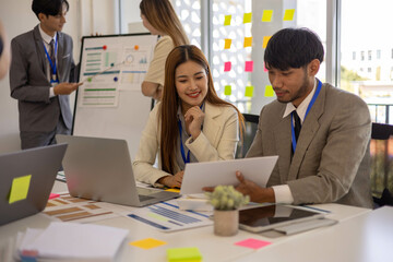 A group of people are working together in a conference room. A man and a woman are looking at a presentation on a laptop. There are several other people in the room
